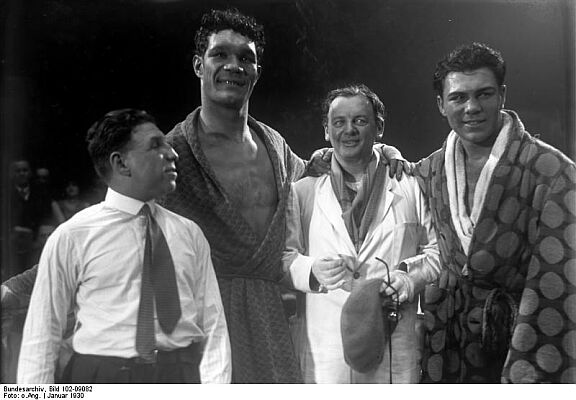 Reinhold Schnzel zwischen den Boxern Max Schmeling (rechts) und Jos Santa bei den Dreharbeiten zum Film "Liebe im Ring"(1930); Quelle: Deutsches Bundesarchiv, Digitale Bilddatenbank, Bild 102-09082; Fotograf: Unbekannt / Datierung: Januar 1930 / Lizenz CC-BY-SA 3.0.