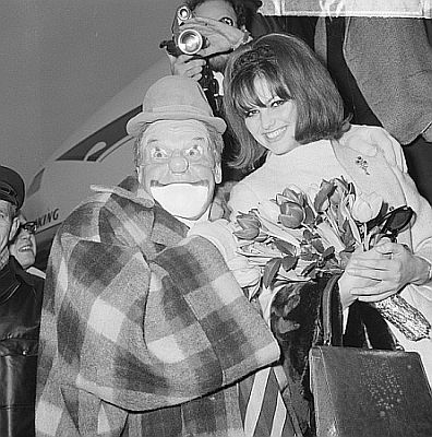 Claudia Cardinale Mitte Dezember 1964 am Flughafen Schiphol mit dem Clown Fantasio; Rechteinhaber: Nationaal Archief (Den Haag, Rjksfotoarchief; Bestandsnummer: 917-2476); Urheber/Fotograf: Nijs, Jac. de / Anefo; Datierung:  16.12.1964; Quelle: Wikimedia Commons; Lizenz: CC0 1.0 (Verzicht auf das Copyright)